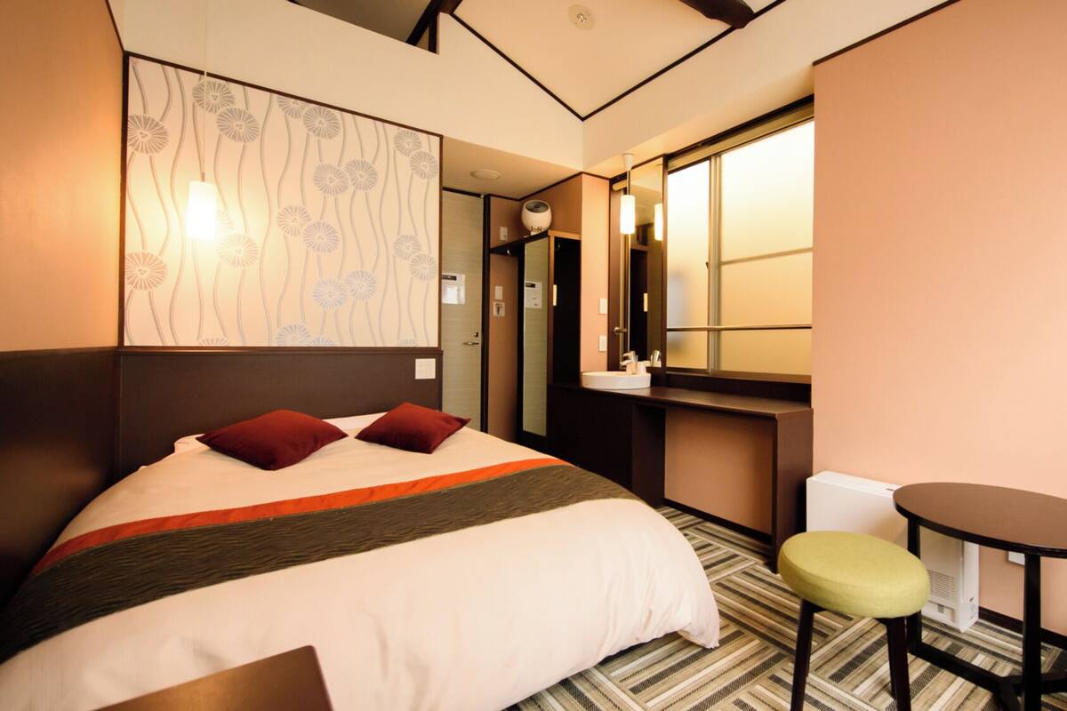 A hotel room with a bed, a small table, a chair, a mirror, and a window.