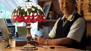 Un homme est assis à un bureau en bois, devant une lampe en vitrail, un ordinateur portable et divers accessoires de bureau.