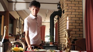 Une personne se tient dans une pièce dont un mur est en briques, avec une table sur laquelle se trouvent des assiettes et une bougie.