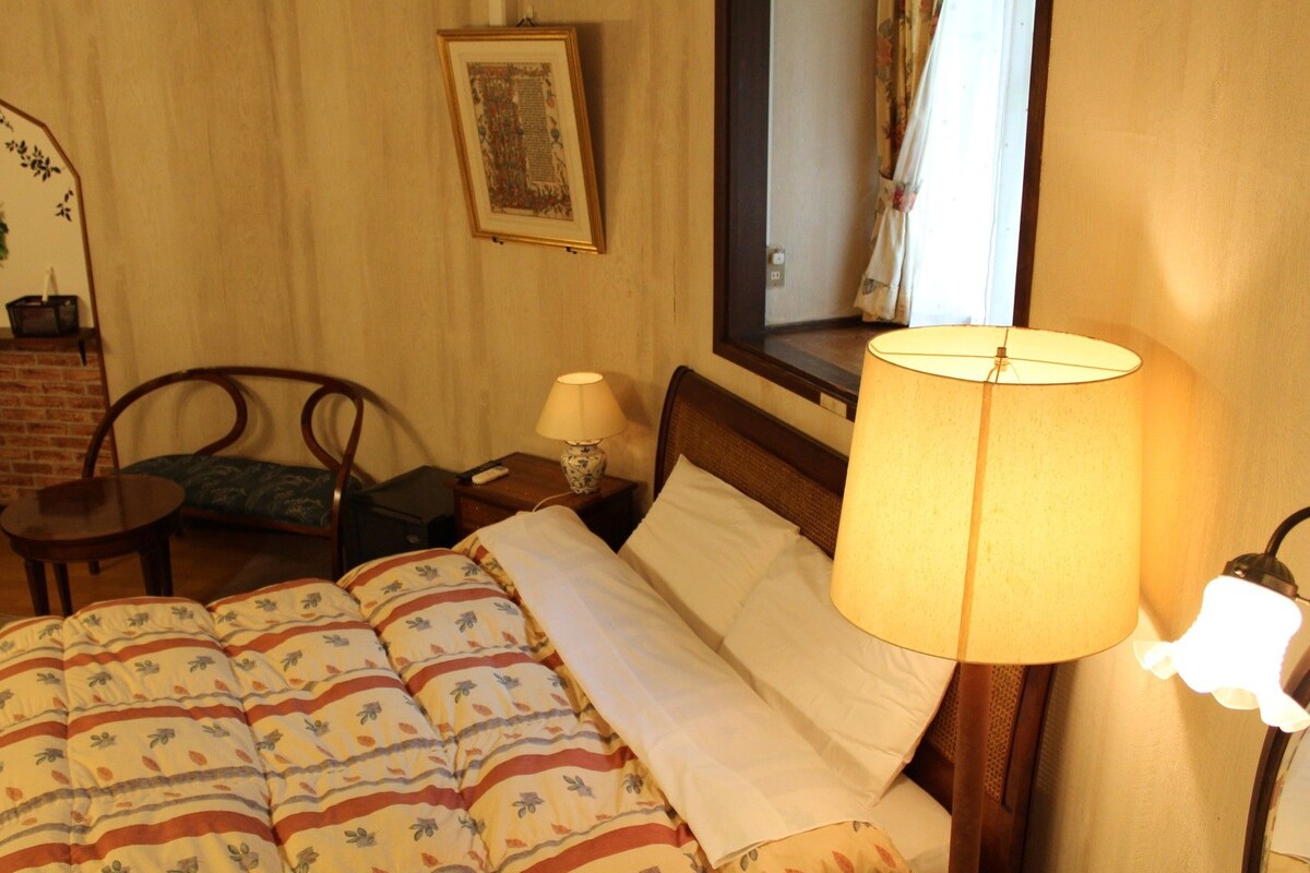 A hotel room with a bed, a nightstand, a lamp, a chair, a small table, a mirror, and a framed picture on the wall.