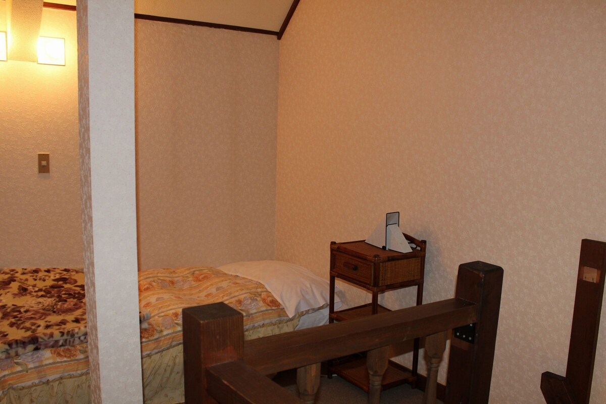 A small, dimly lit room with a single bed, a nightstand with a lamp, and a wooden chair.