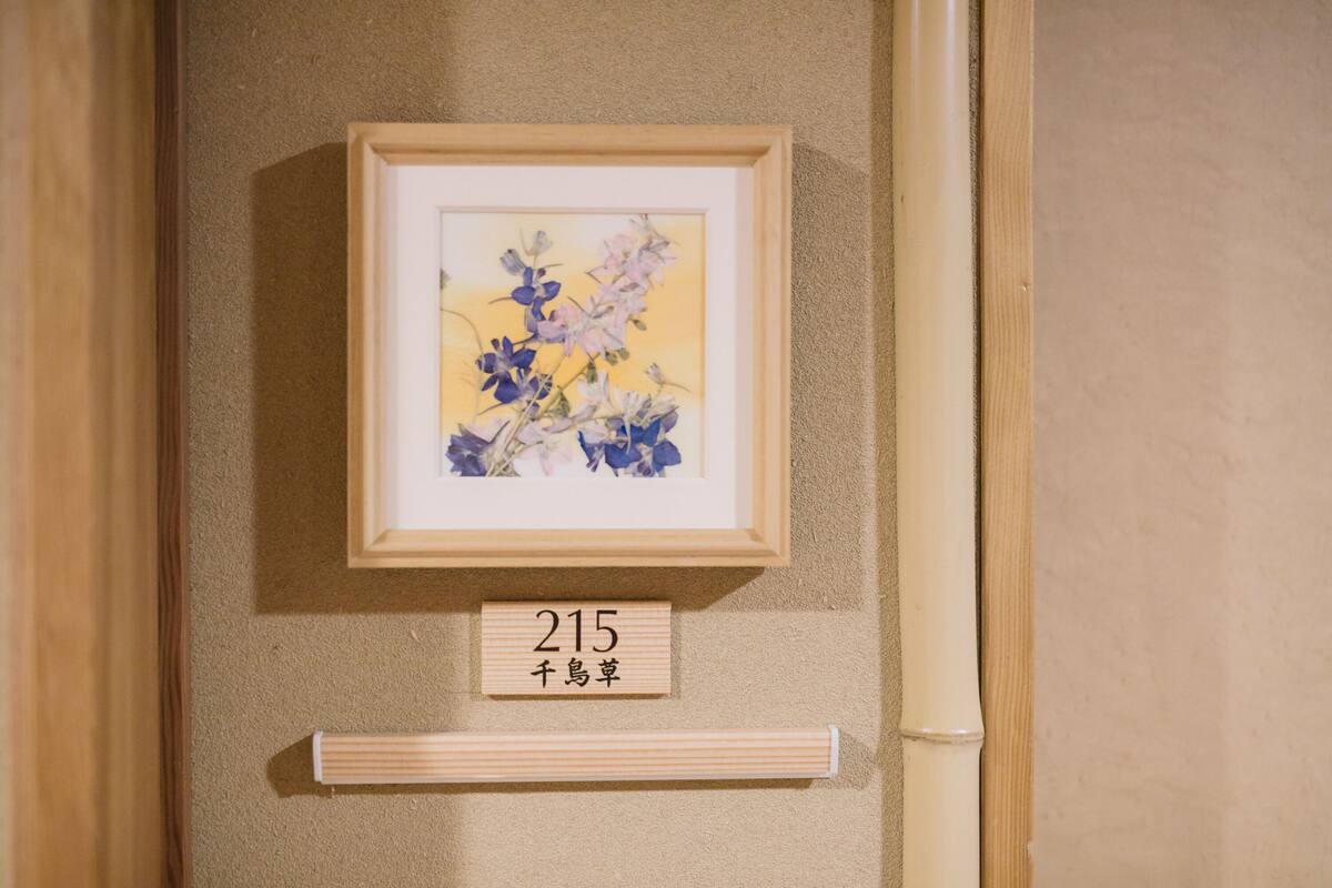 Ein gerahmtes Bild mit Blumen an einer Wand, unterhalb eine Beschilderung mit der Aufschrift '215' und japanischen Schriftzeichen.