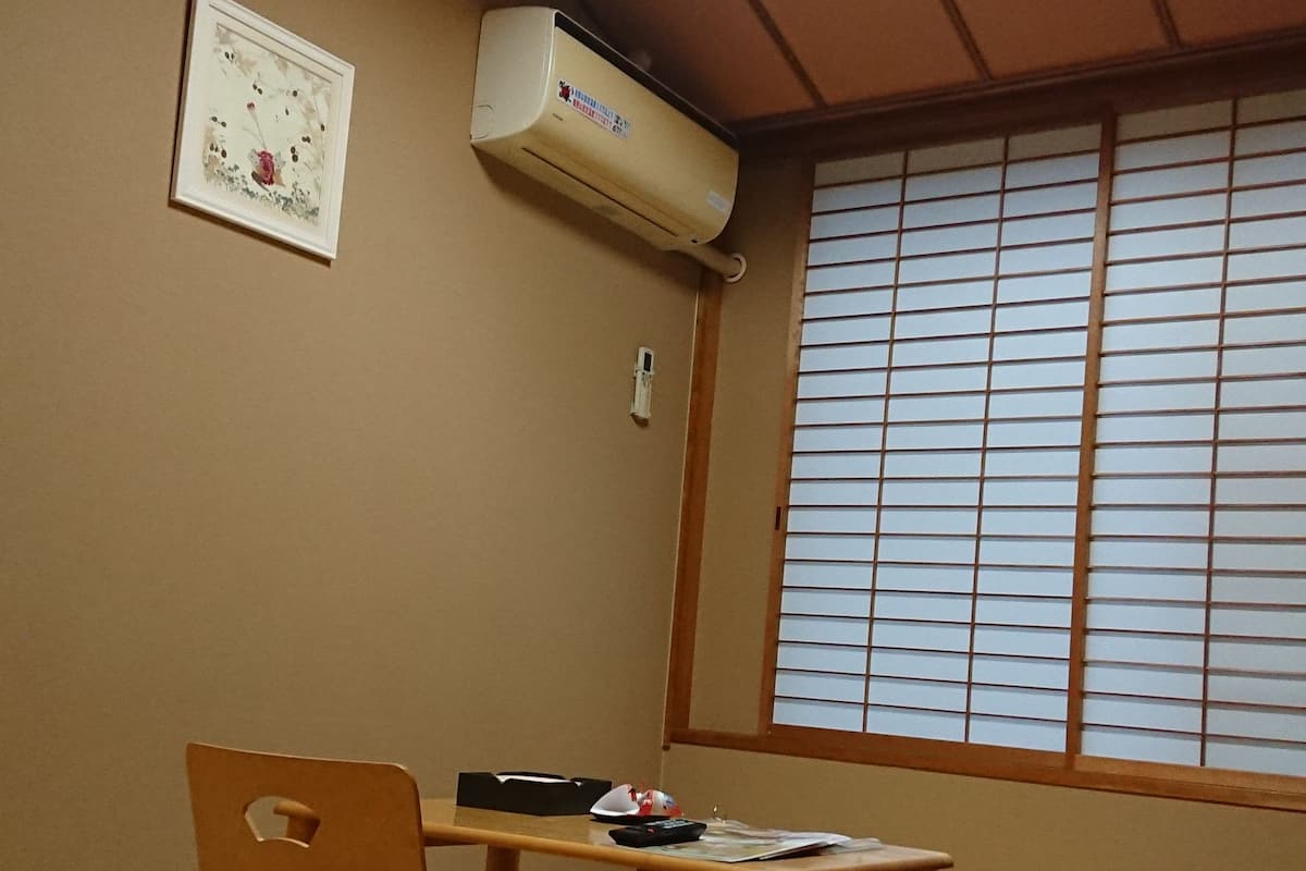 Ein traditioneller japanischer Raum mit Tatami-Bodenbelag, einem niedrigen Holztisch, einem gepolsterten Sessel, einer schwarzen Aufbewahrungsbox, einem gerahmten Bild und einem Fenster mit Shoji-Schirm.