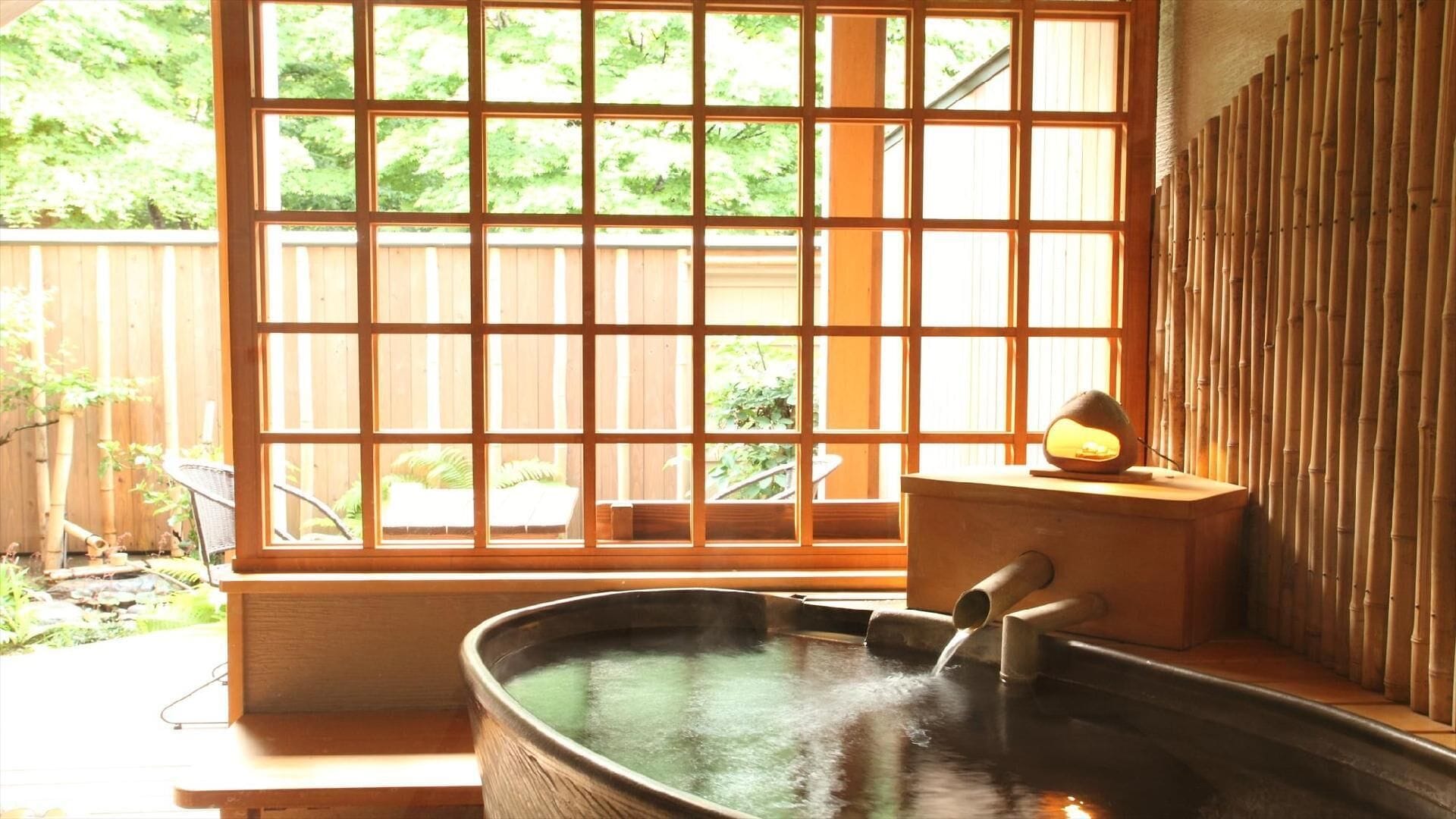 Un onsen japonès tradicional amb una banyera de fusta, paret de bambú i vista a la vegetació a través d'una finestra de reixeta.