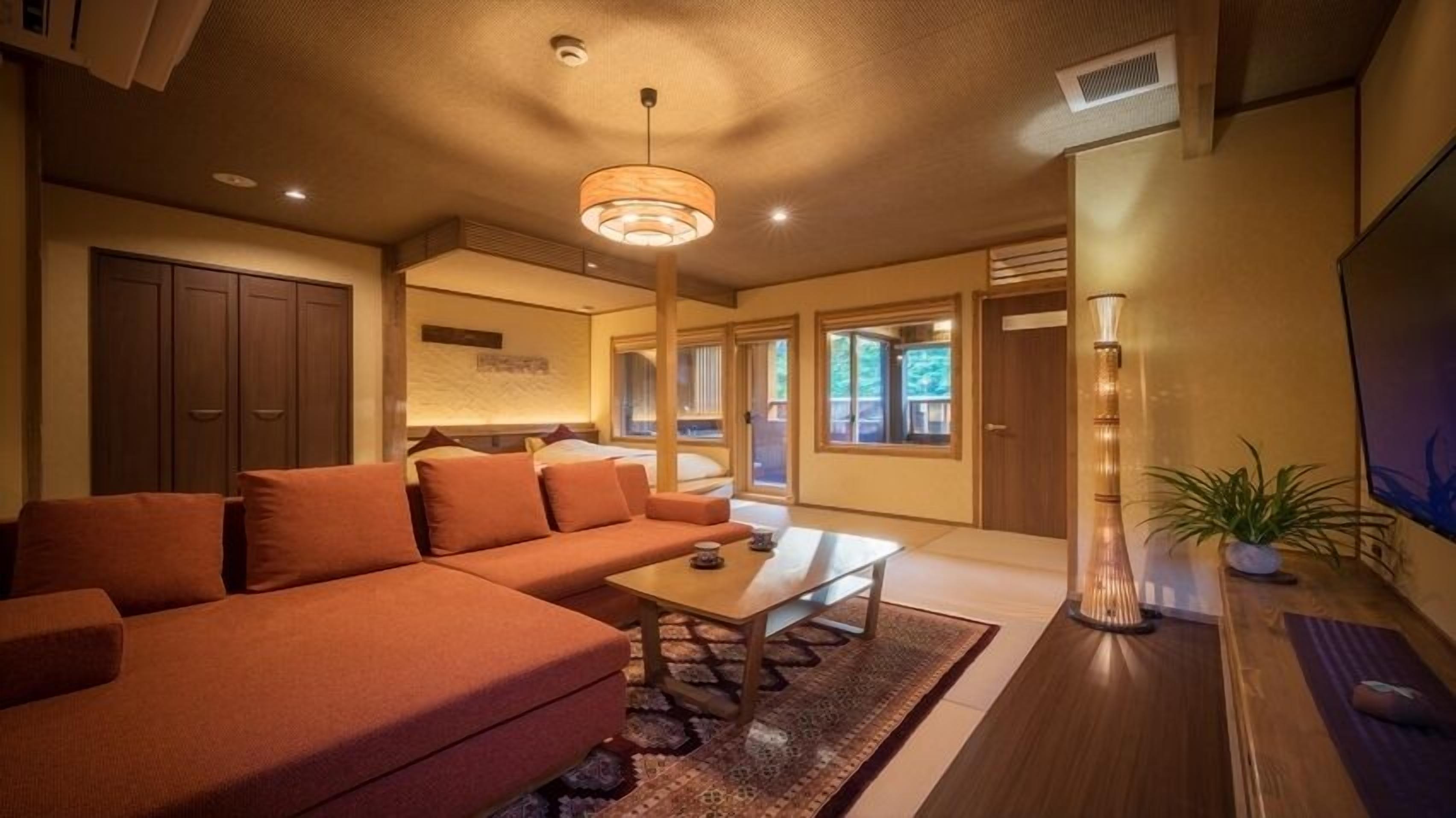 Ein modernes Wohnzimmer mit einem Ecksofa, einem Couchtisch und einem Fernseher. Es gibt Schiebetüren, die auf einen Balkon führen.