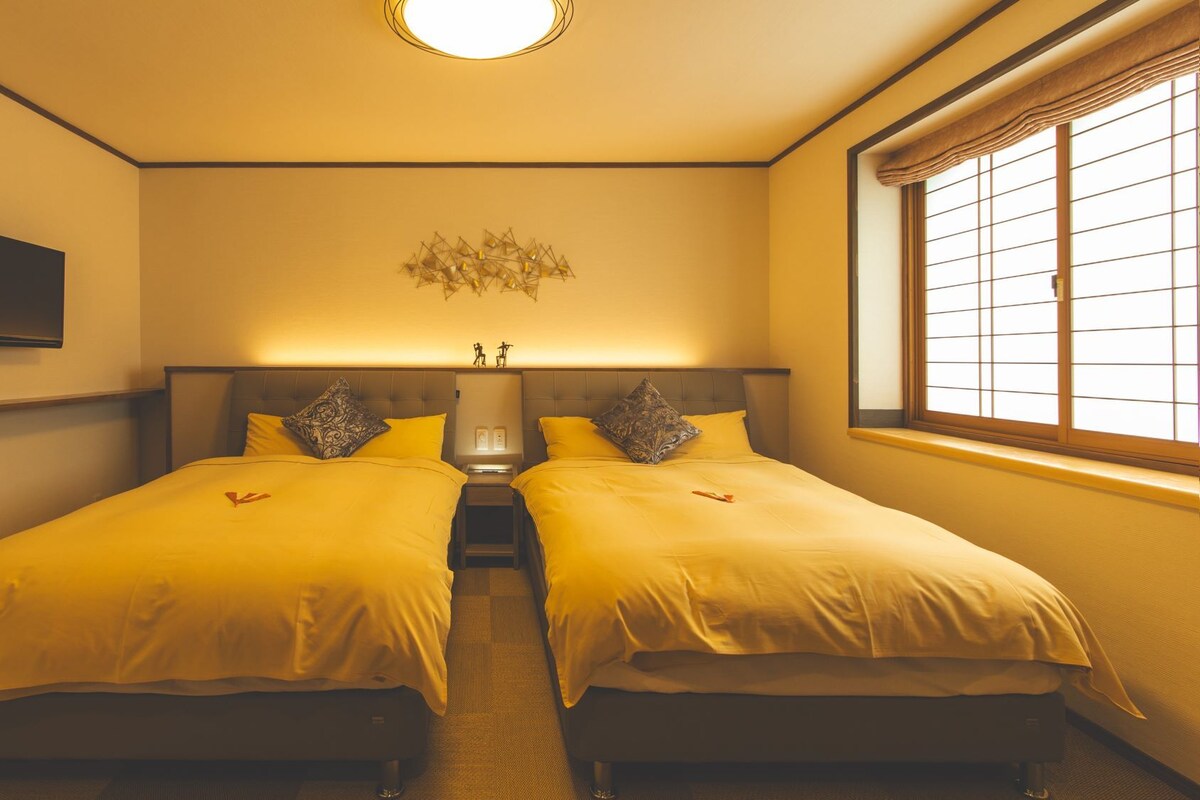 A hotel room with two beds, a flat-screen TV, a decorative wall piece, and a window with blinds.