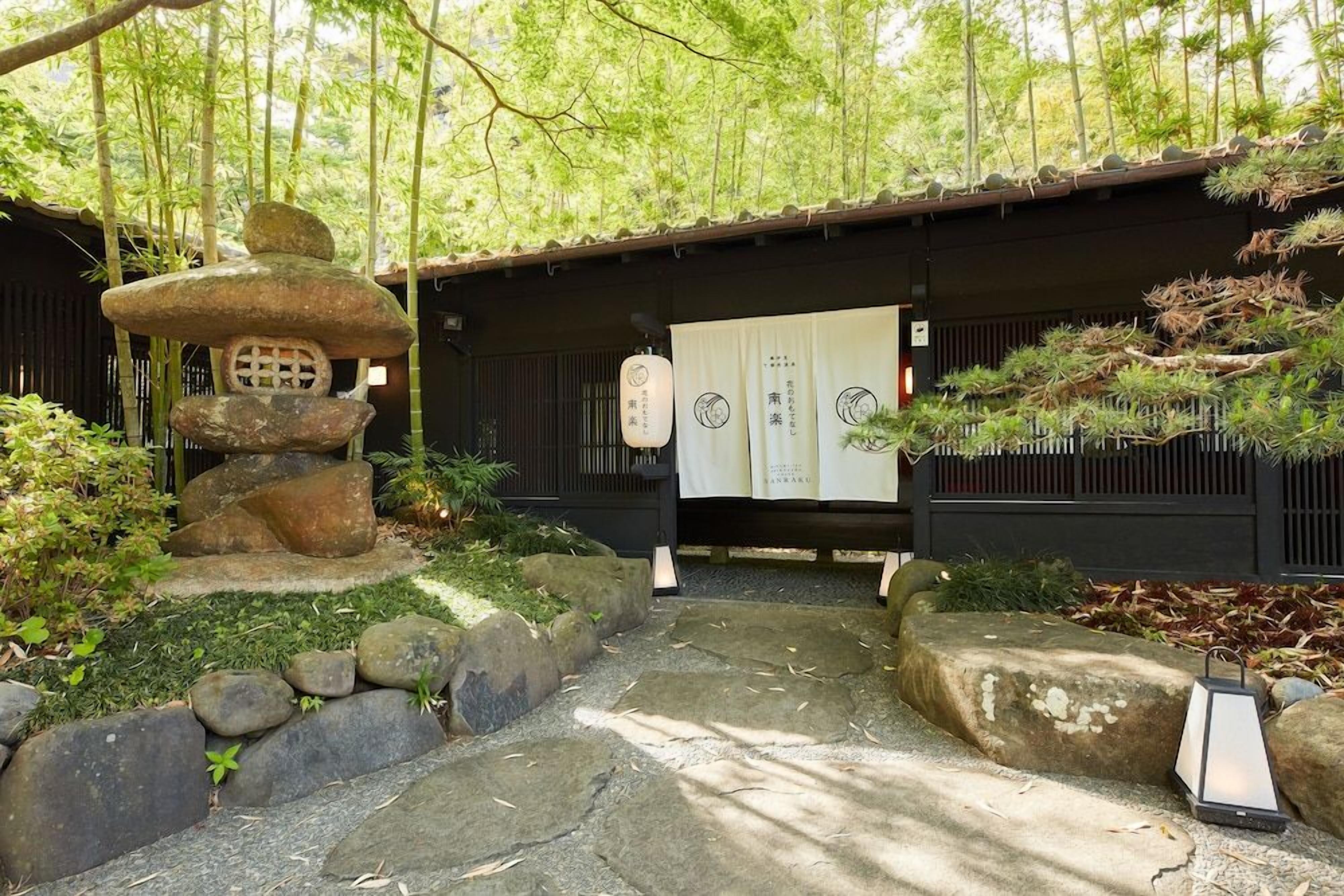 A traditional Japanese garden with a stone lantern, bamboo, and a wooden building with sliding doors.