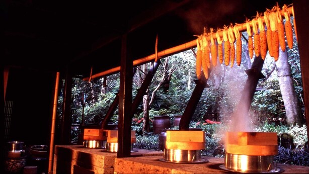 A traditional cooking setup with hanging corn cobs and steaming pots, set in a shaded outdoor area with a view of greenery. - Hana No Omotenashi Nanraku (Minamiizu)