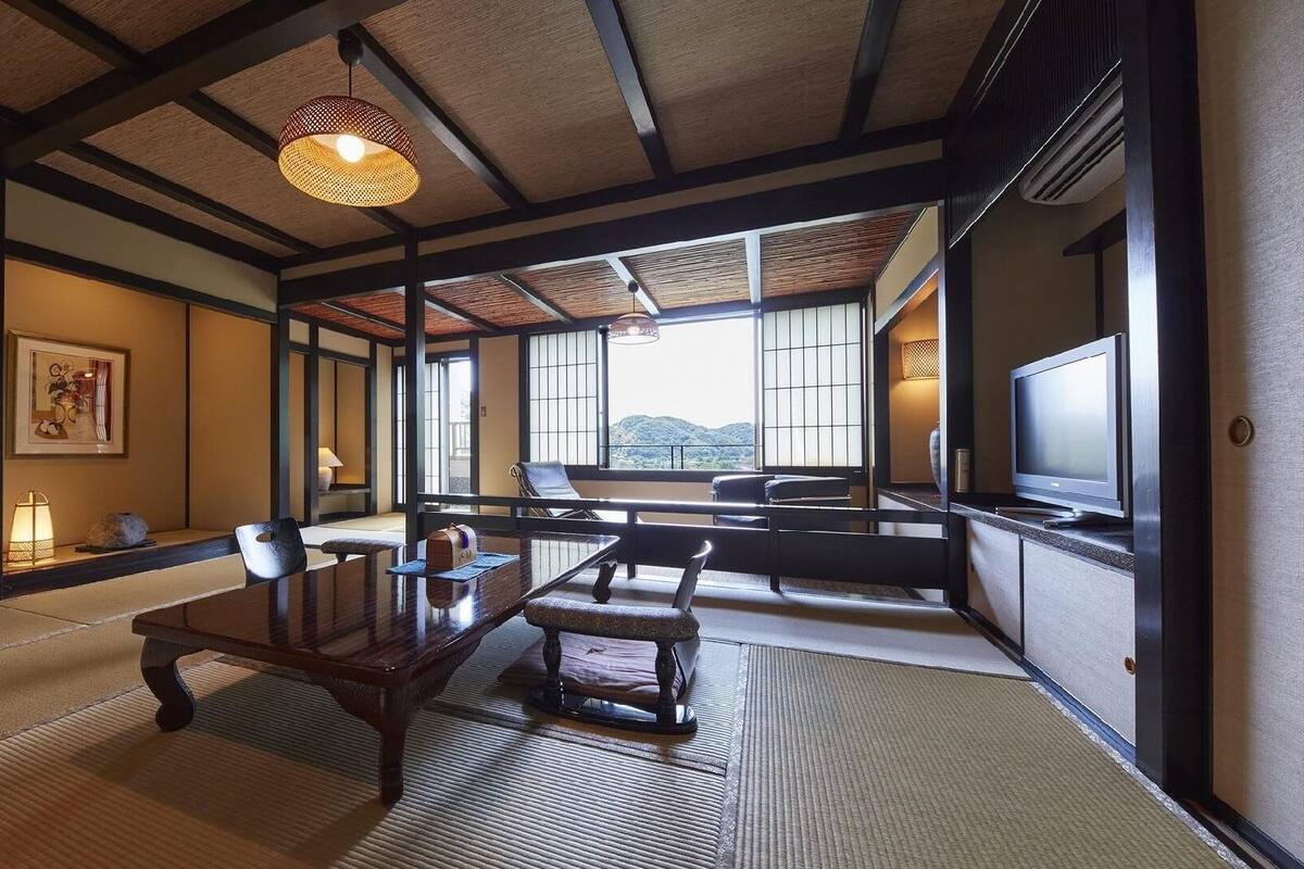 Ein traditioneller japanischer Raum mit Tatami-Boden, einem niedrigen Tisch, Stühlen, einem Fernseher und großen Fenstern mit Blick ins Freie.