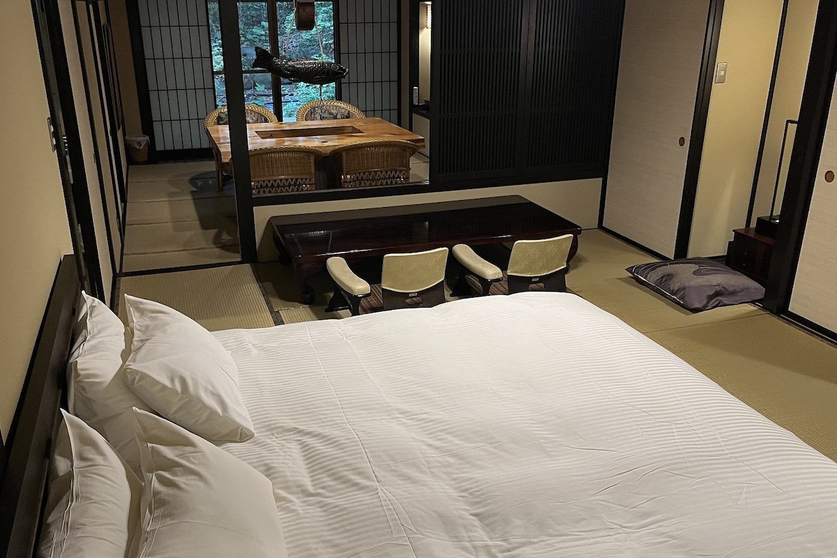 A hotel room with a bed, a small table, two chairs, a sofa, and a view of a forest through a window.