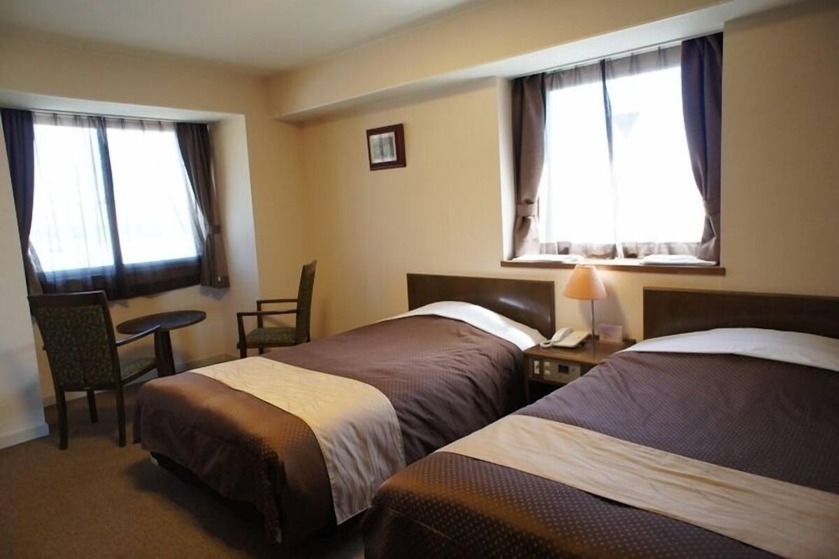 A hotel room with two beds, a desk, a chair, and a lamp.