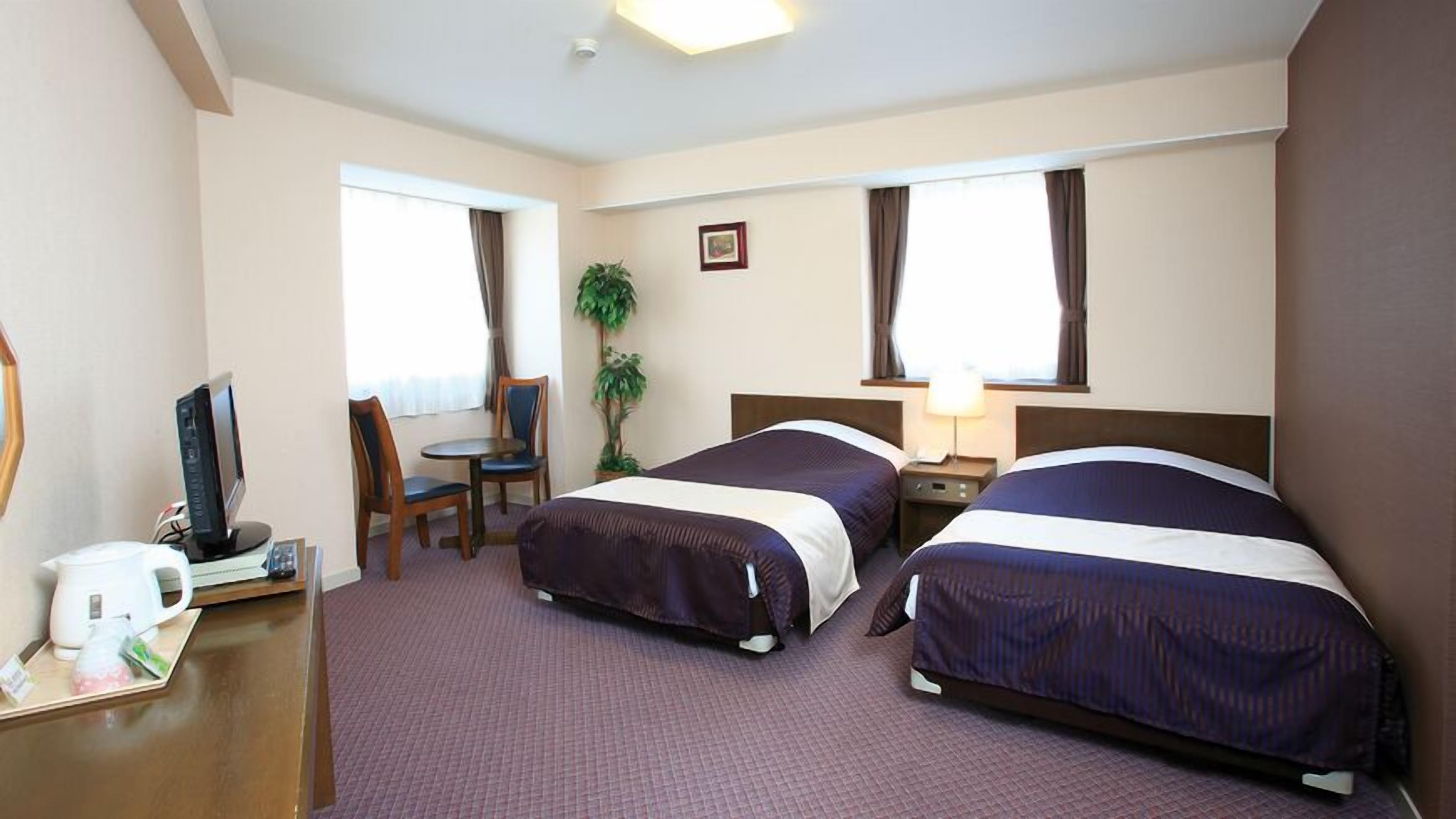A hotel room with two beds, a desk with a computer, a chair, a plant, and a window.