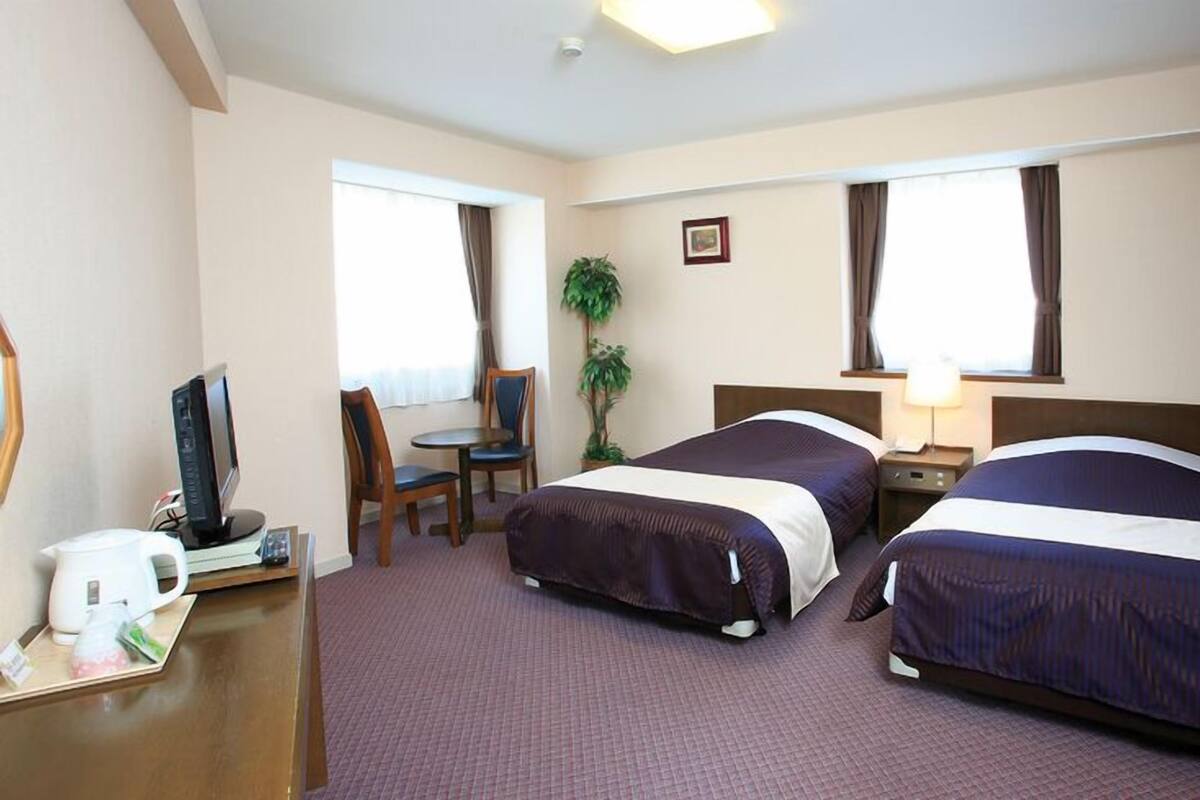 A hotel room with two beds, a desk with a computer, a chair, a plant, and a window.