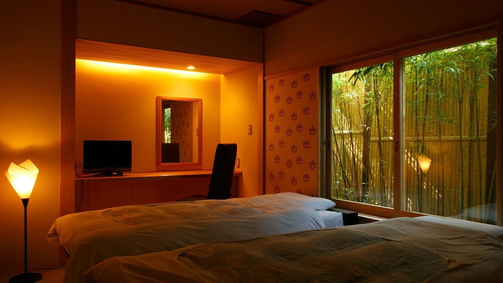 A hotel room with two beds, a desk with a computer, a chair, and a large window with bamboo outside.