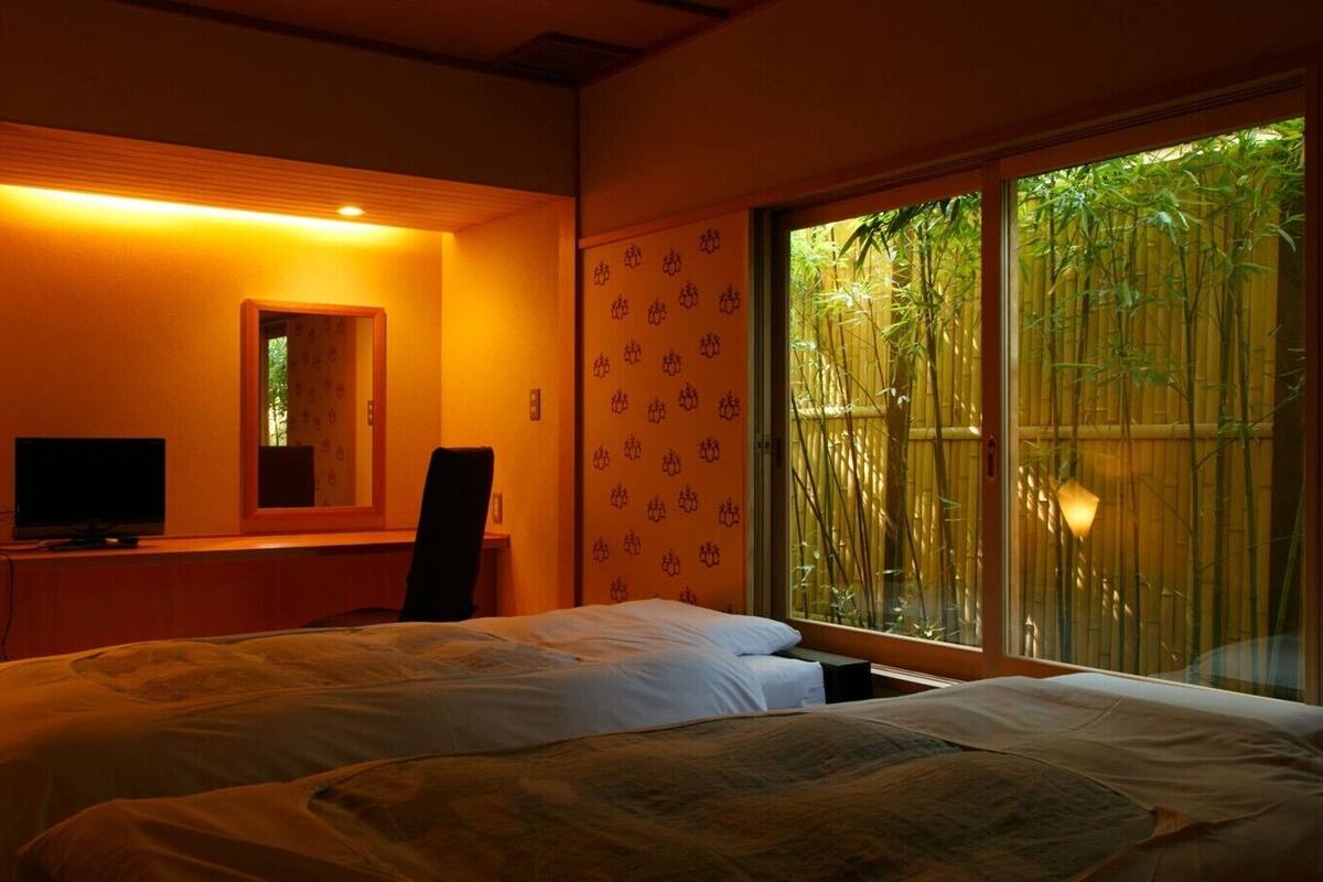 A hotel room with two beds, a desk with a computer, a chair, and a large window with bamboo outside.