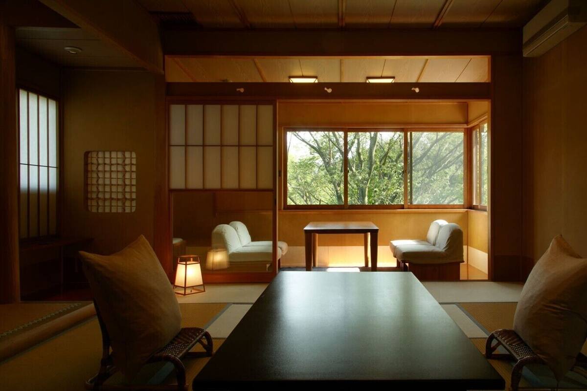 A traditional Japanese room with tatami flooring, a low table, two chairs, and a large window with a view of trees.