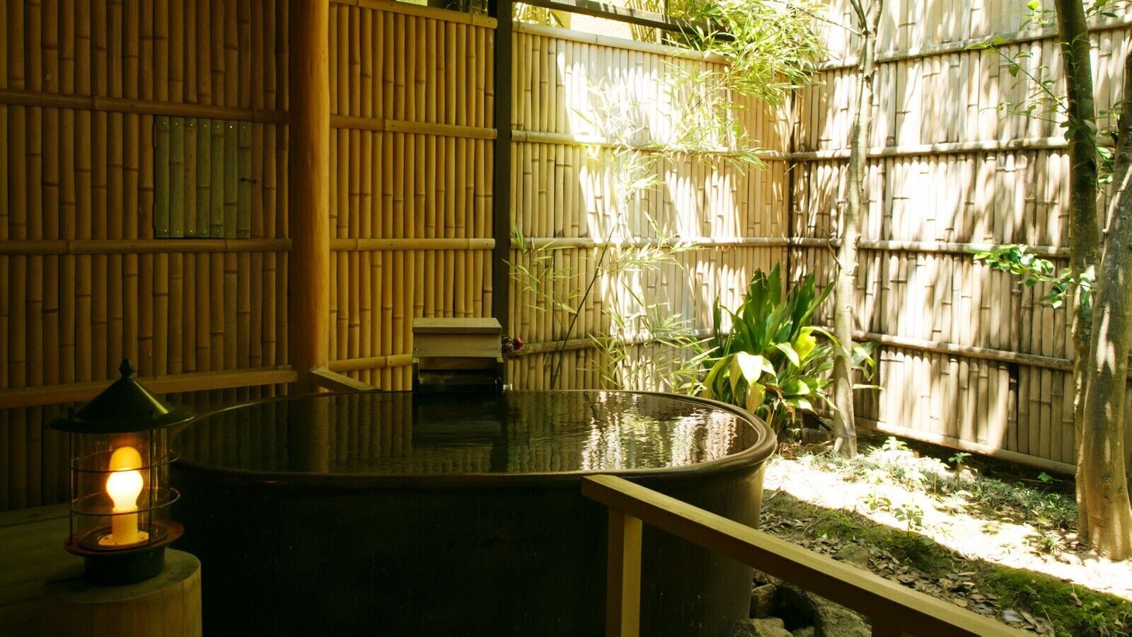 A tranquil outdoor area with a water feature, bamboo walls, and a small table with a lantern.