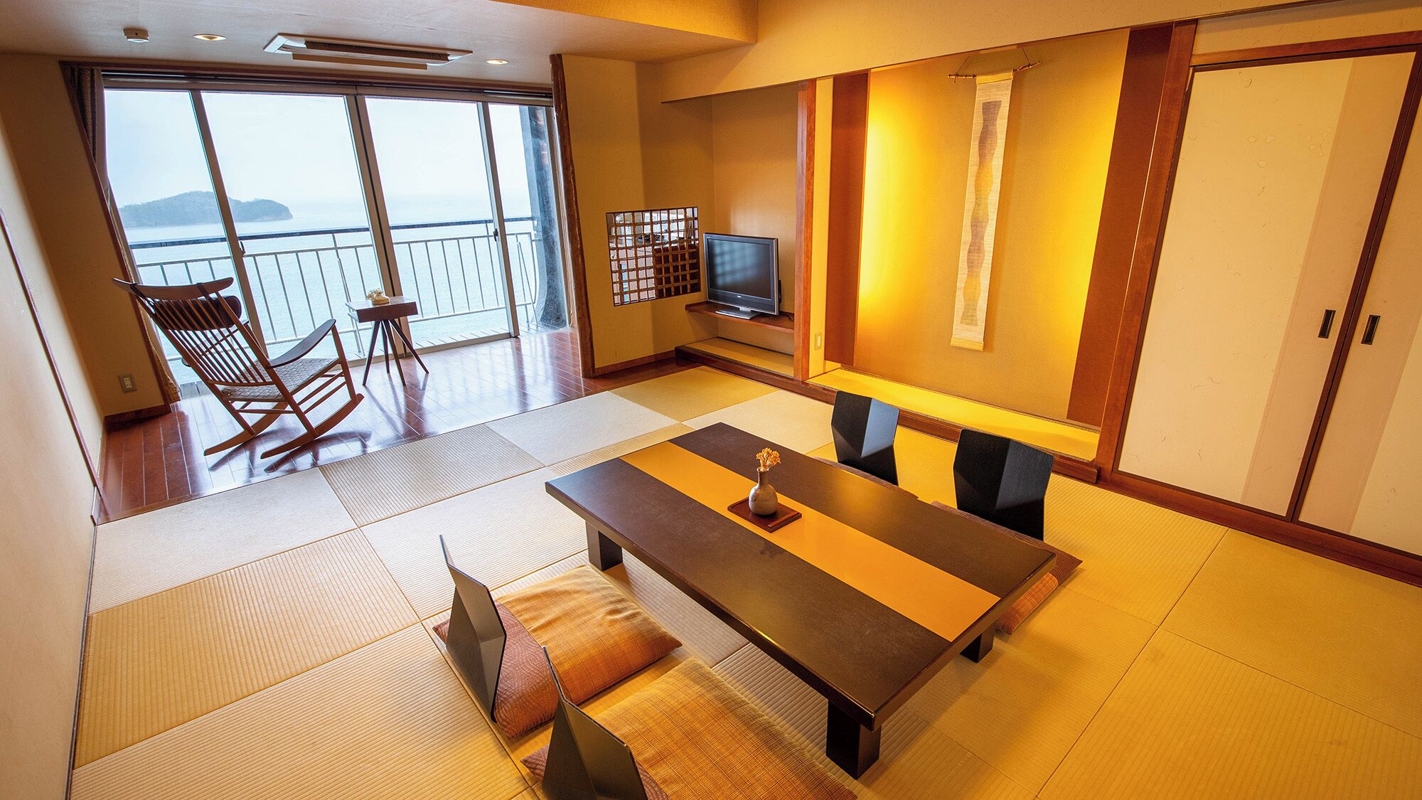 Una habitación con suelo de tatami, una mesita baja con cojines, un televisor y una puerta corredera que da a un balcón con vistas al mar.