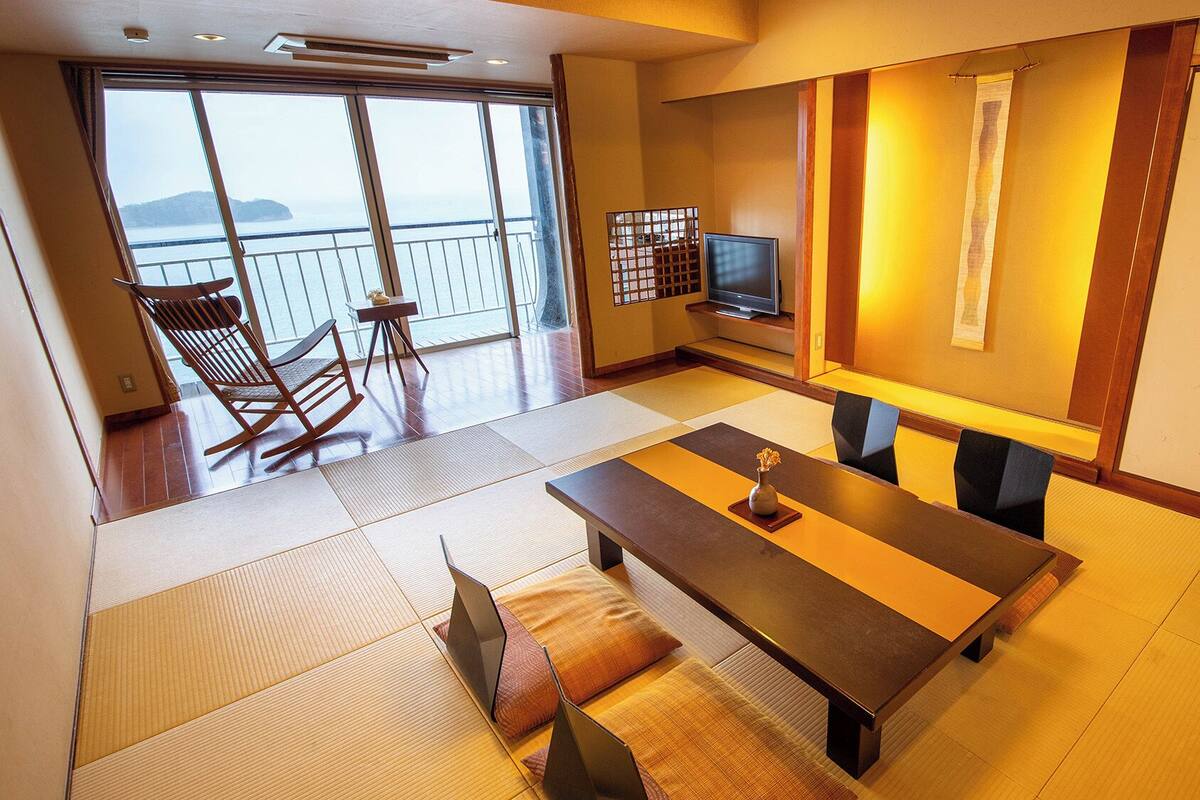 A room with tatami flooring, a low table with cushions, a television, and a sliding door leading to a balcony with a view of the sea.