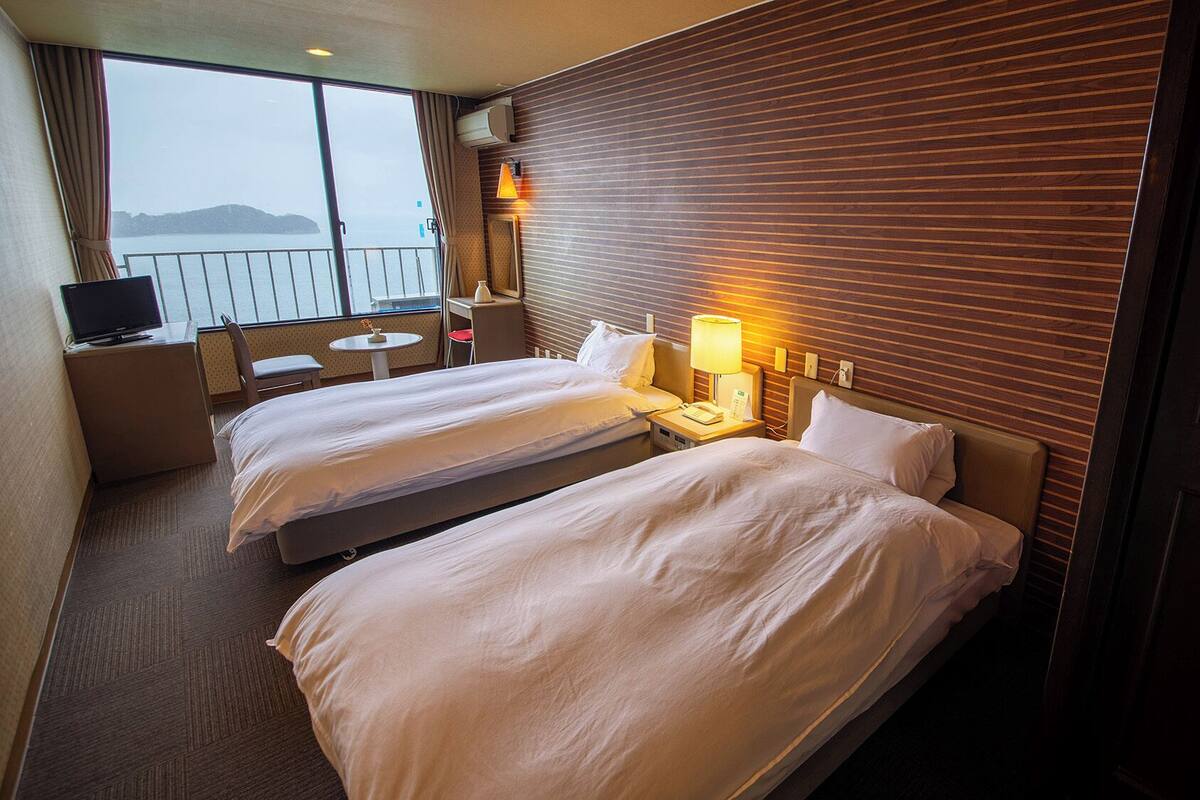 A hotel room with two beds, a desk, a chair, a small table, and a view of the sea.
