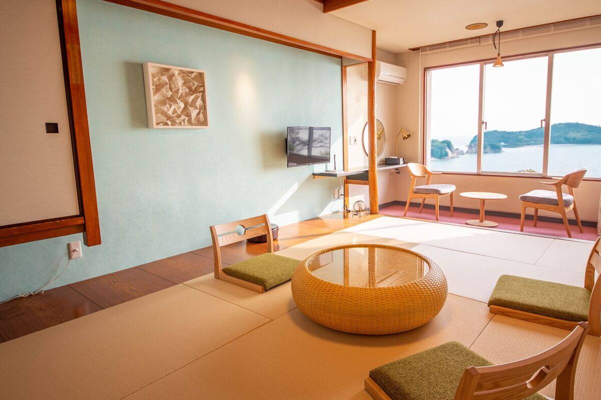 A room with tatami flooring, a low table with cushions, a woven round chair, and a view of the sea through large windows.