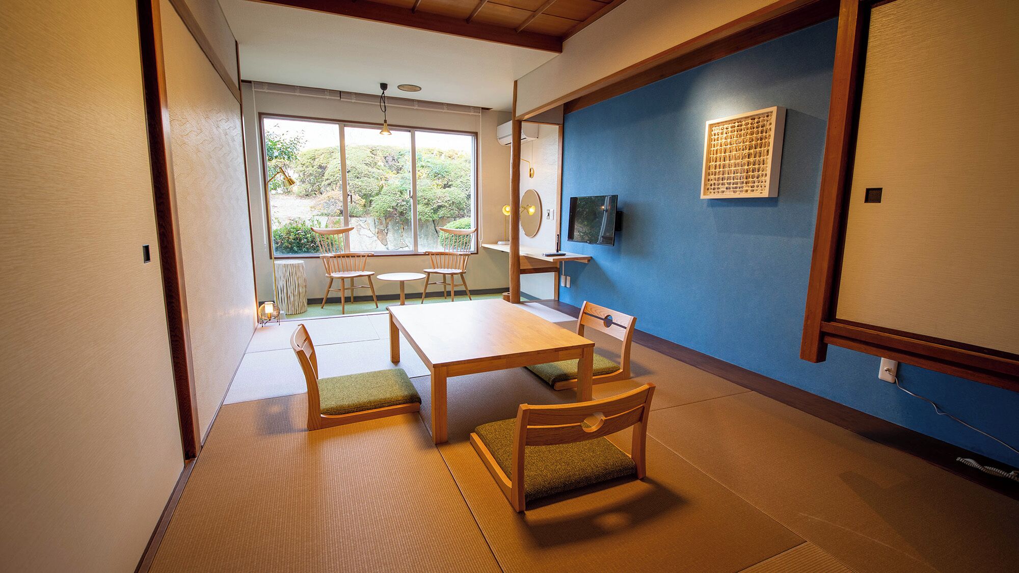 Una habitación japonesa tradicional con suelo de tatami, una mesa baja, sillas, un televisor y una ventana con vistas a la vegetación.