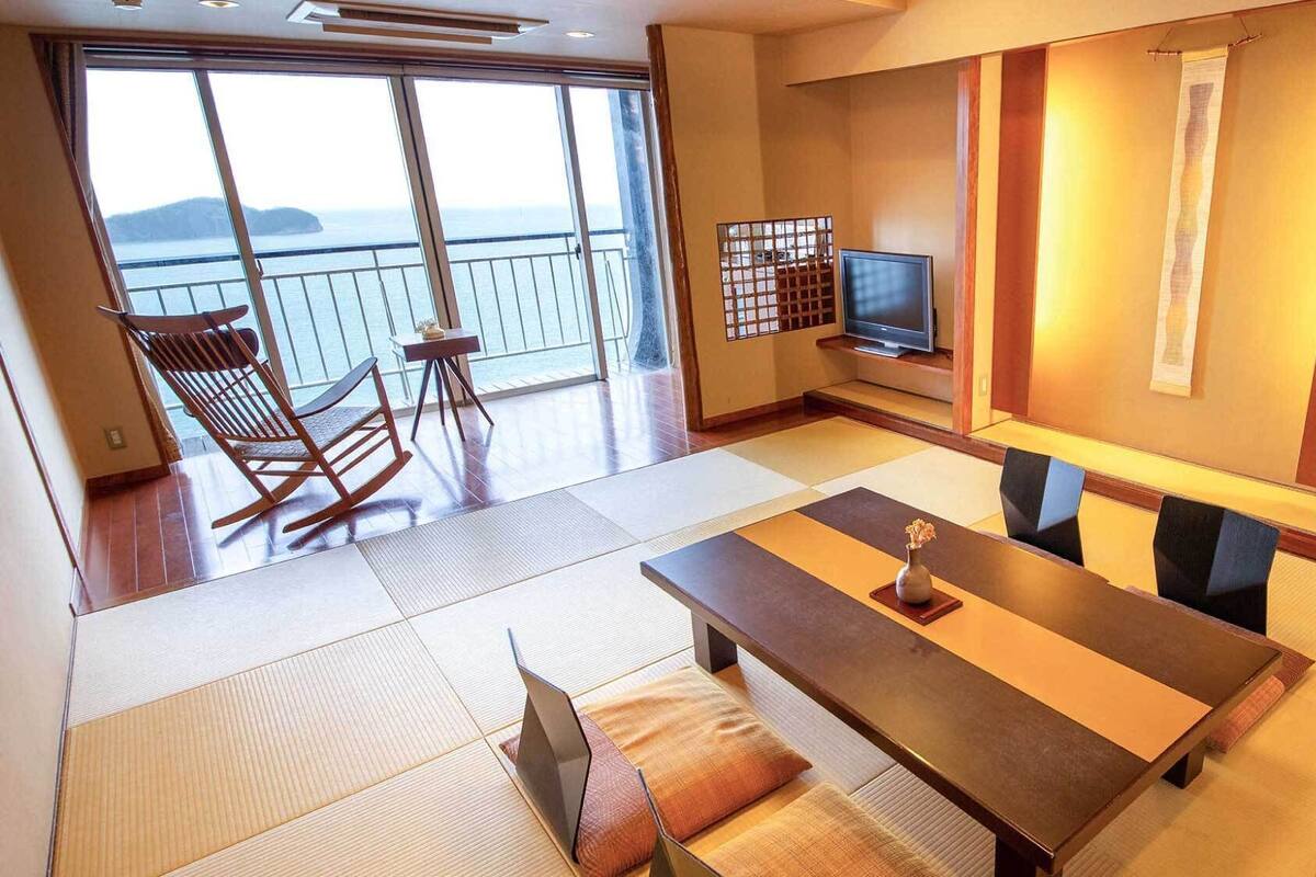 A room with a large window, a television, a dining table, and a seating area with chairs.