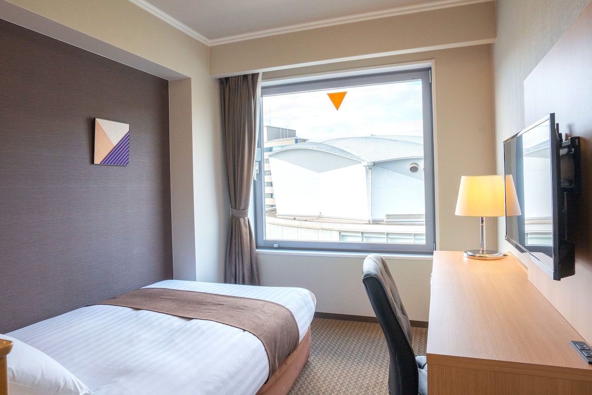 Habitación de hotel con cama, escritorio con lámpara, televisor y ventana con vistas a edificios.