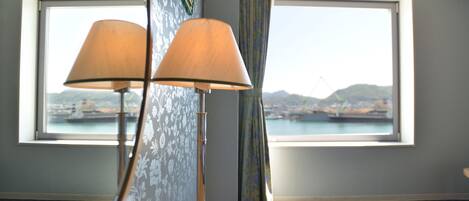A room with a view of a harbor, a table with a lamp, and patterned wallpaper.