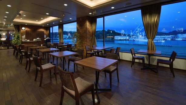 Um restaurante com mesas e cadeiras de madeira, grandes janelas com vista para um porto e navios, e um relógio na parede.