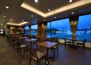 A restaurant with wooden tables and chairs, large windows with a view of a harbor and ships, and a clock on the wall. - Green Hill Hotel Onomichi (Onomichi)