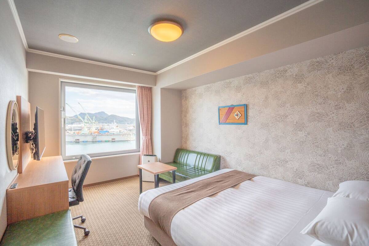A hotel room with a bed, a desk with a chair, a small table, a window with a view of a harbor, and a decorative wall piece.