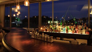 Um bar com balcão curvo, luminárias pendentes e vista para a paisagem urbana noturna.