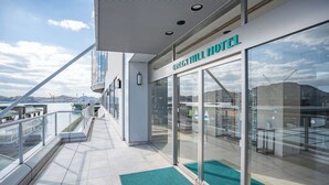 Um hotel moderno com fachada de vidro, um tapete de entrada verde e vista para a água e a paisagem urbana.