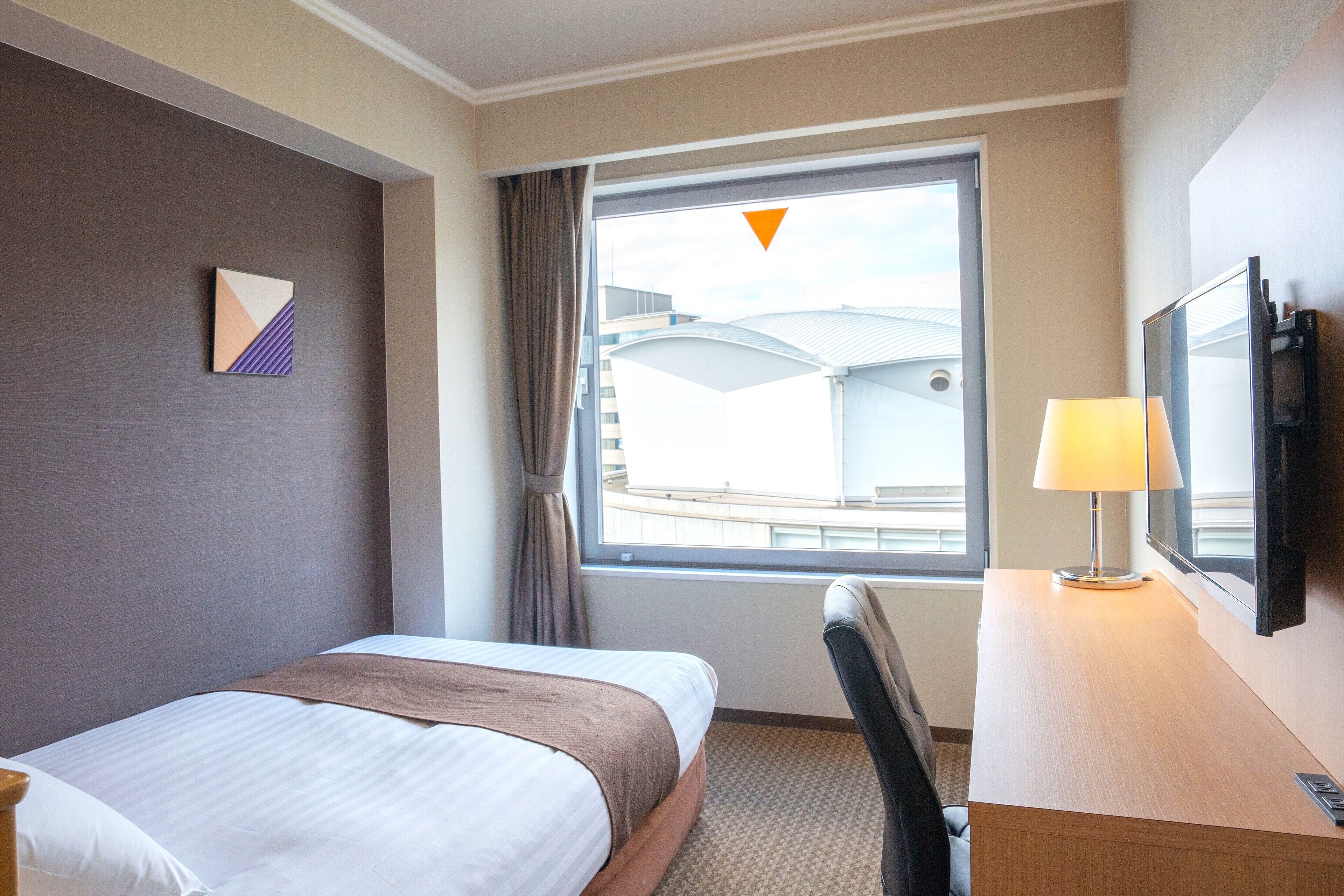 Habitación de hotel con cama, escritorio con lámpara, televisor y ventana con vistas a edificios.