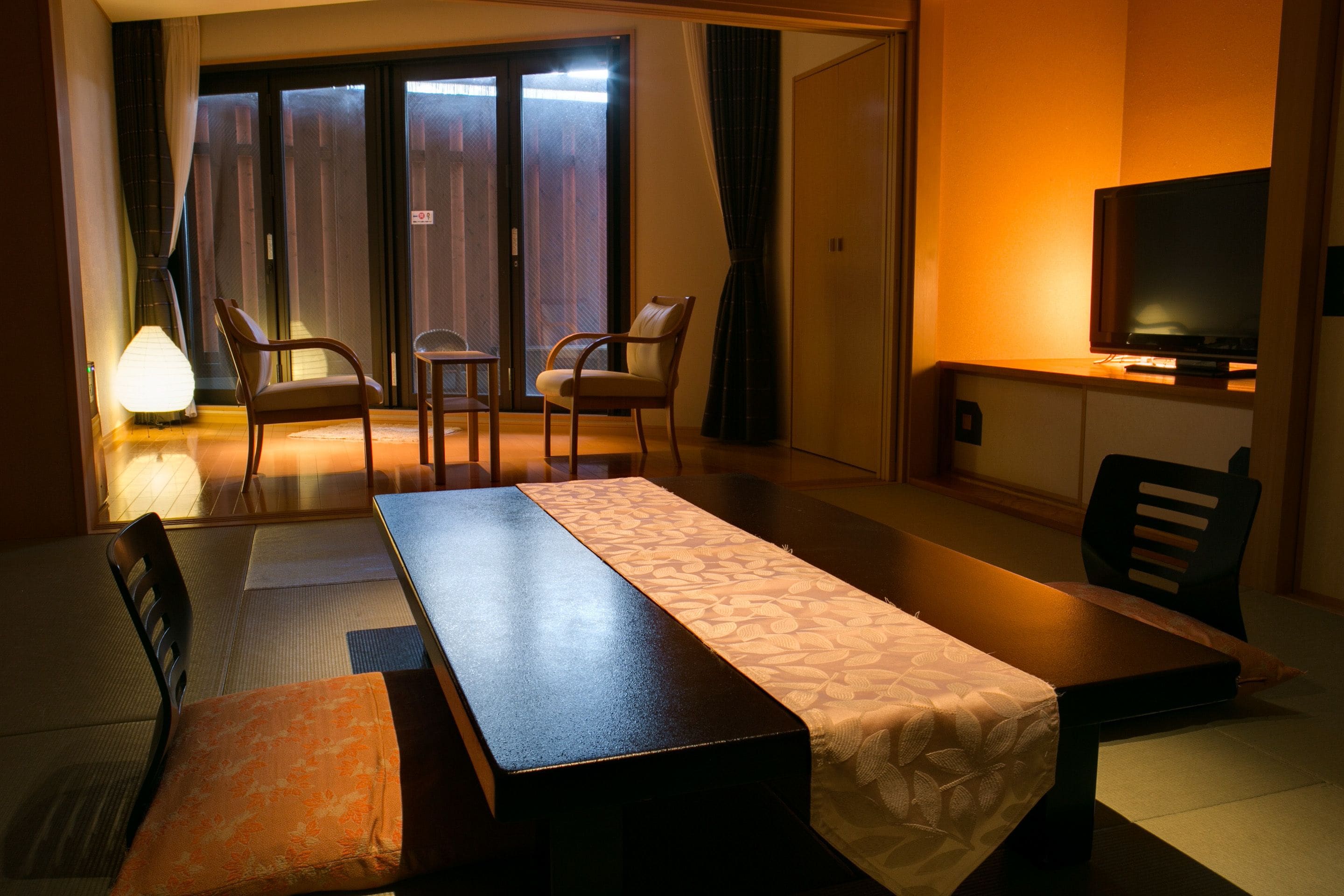 Habitación de hotel con cama, escritorio, dos sillas, televisor y puerta corrediza a un balcón.