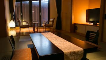 Habitación de hotel con cama, escritorio, dos sillas, televisor y puerta corrediza a un balcón.