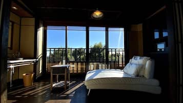 A room with a bed, a small table, a chair, and a view through large windows.