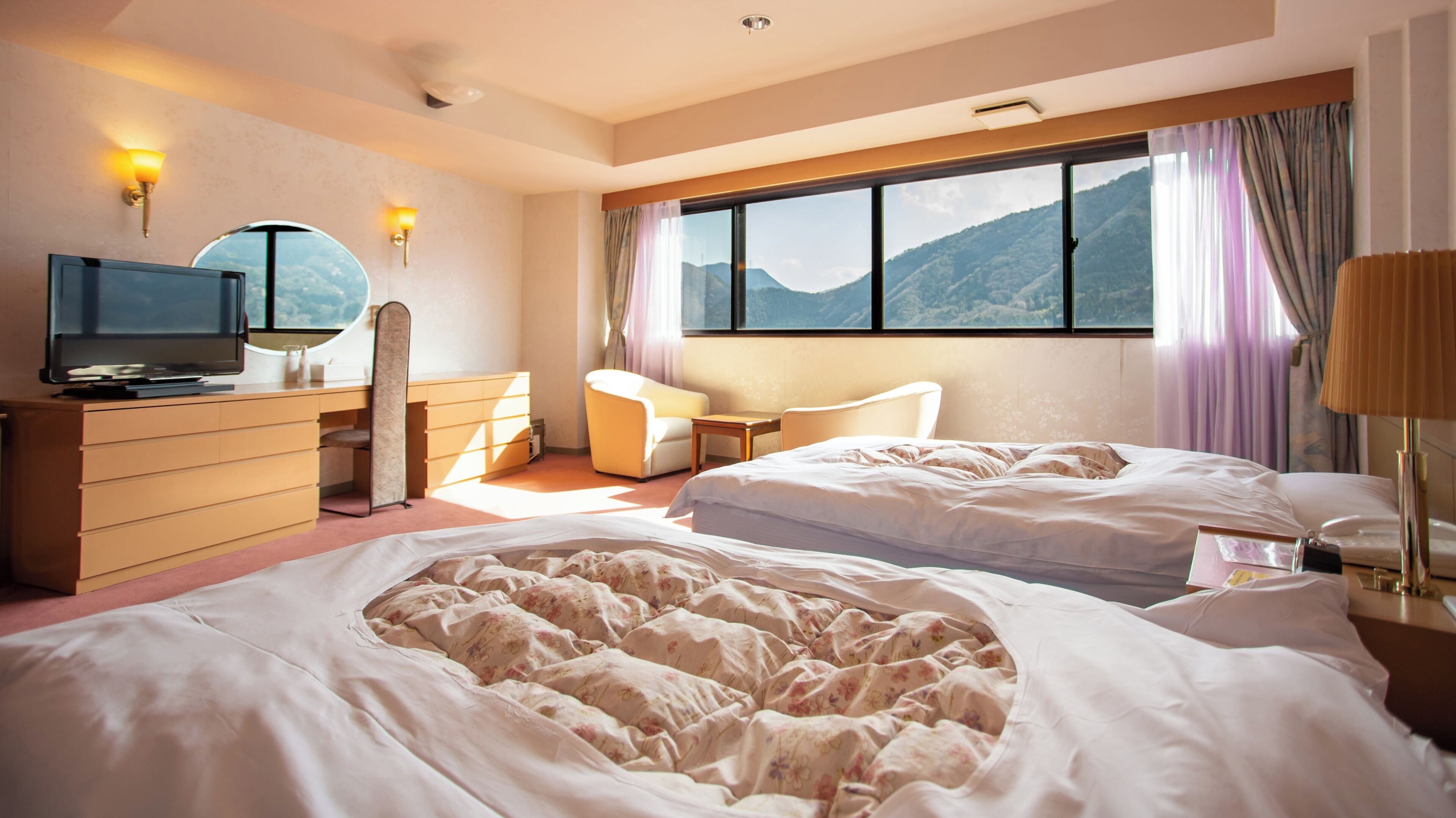 Habitación de hotel con dos camas, un escritorio, una silla, un televisor y ventana con vista a la montaña.