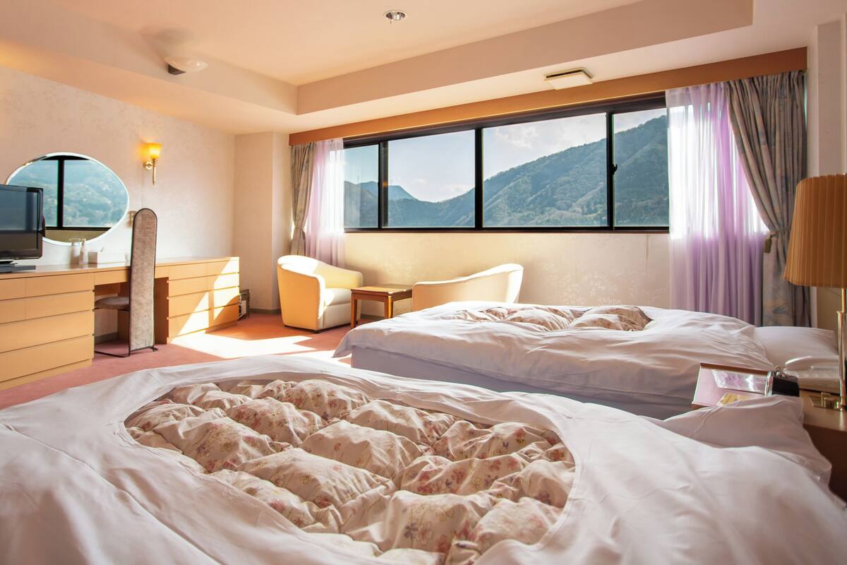 Habitación de hotel con dos camas, un escritorio, una silla, un televisor y ventana con vista a la montaña.
