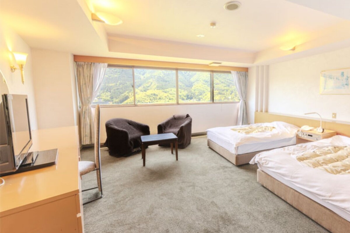 Une chambre d’hôtel avec deux lits, un bureau, une chaise et une grande fenêtre donnant sur l’extérieur.