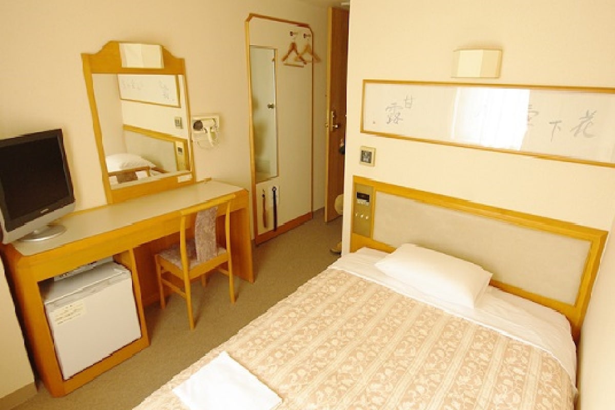 A hotel room with a bed, a desk with a computer, a chair, a mirror, and a wardrobe.