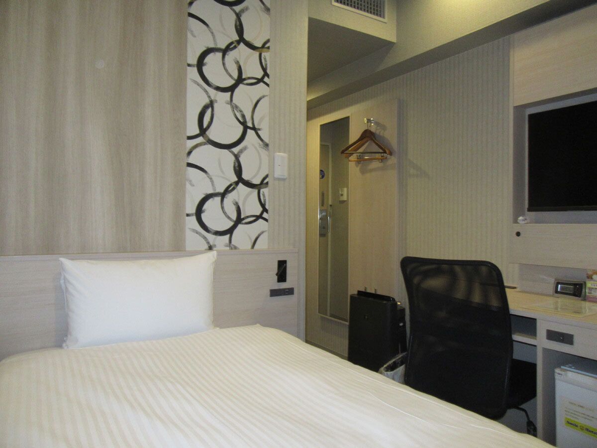 Una habitación de hotel con cama, escritorio, silla, perchero y televisor.
