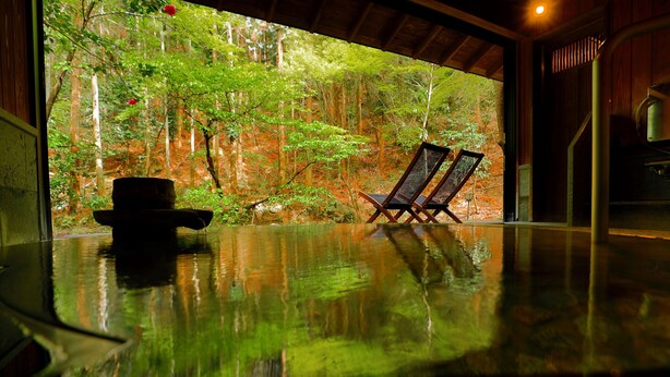 Una habitación con piso reflectante, dos sillas, una mesa y vista a un bosque a través de un ventanal grande.