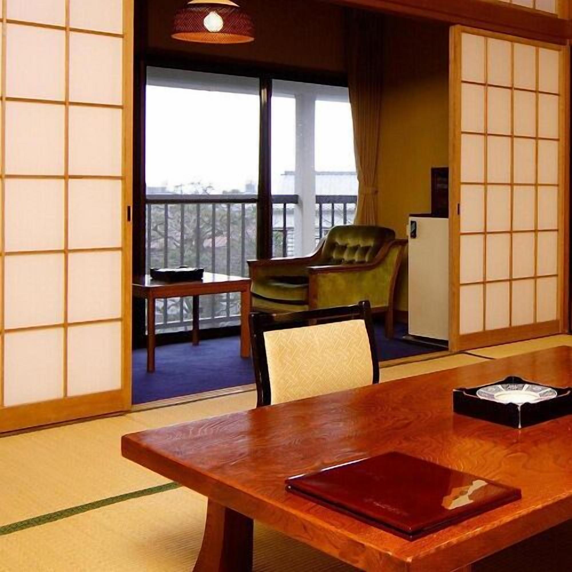 Una habitación tradicional japonesa con suelo de tatami, una mesa de madera con una carpeta roja, una silla verde y vistas a un balcón a través de una puerta corredera.
