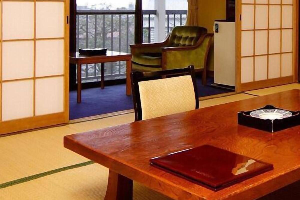 Ein traditioneller japanischer Raum mit Tatami-Bodenbelag, einem Holztisch mit einer roten Mappe, einem grünen Stuhl und Blick durch eine Schiebetür auf einen Balkon.