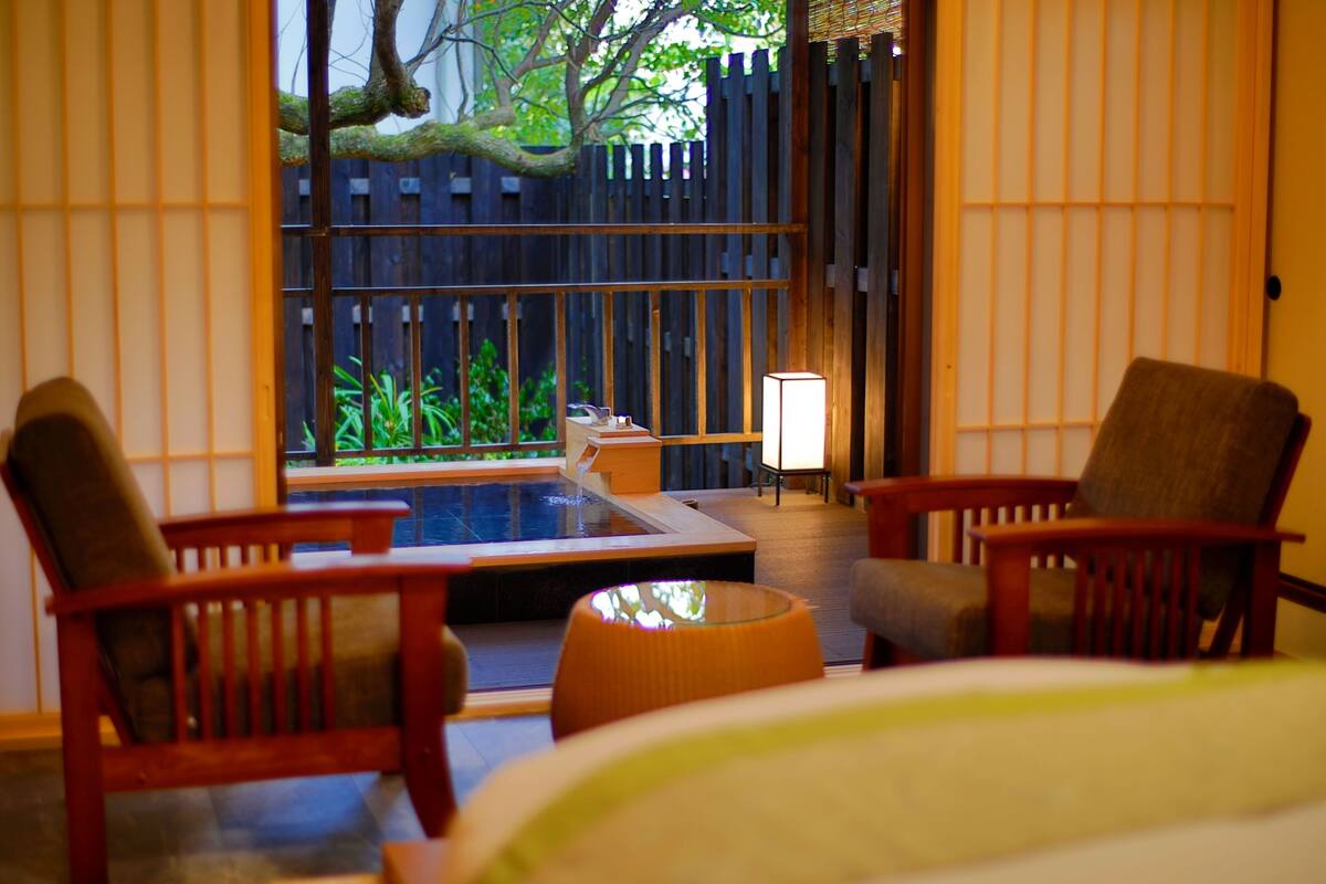 Ein Zimmer mit einem Bett, zwei Holzstühlen, einem kleinen Tisch und Blick auf einen Garten mit Holzzaun und Pflanzen.
