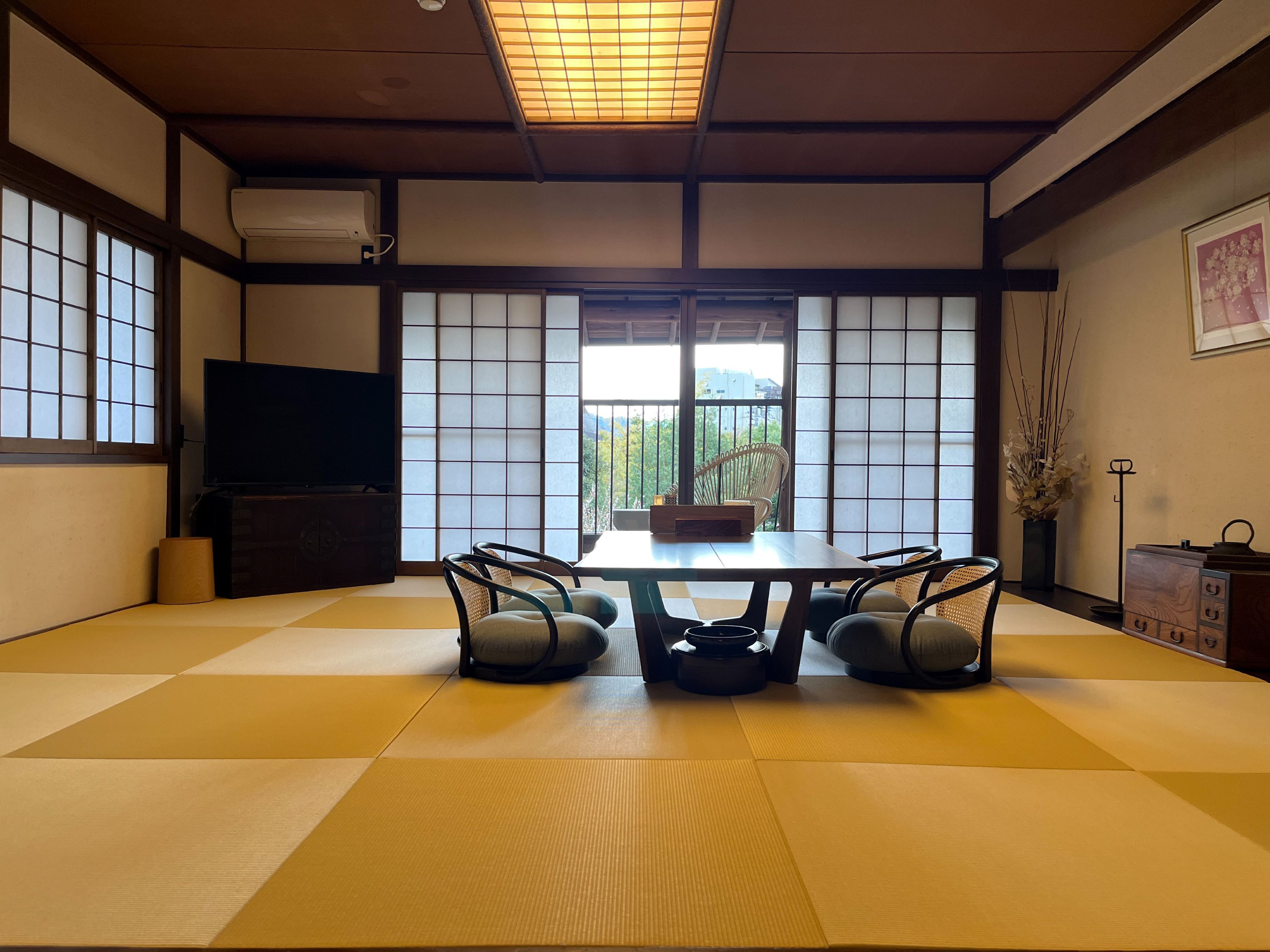 A tatami mat room with a low table and four chairs, a television, and a view of greenery outside.