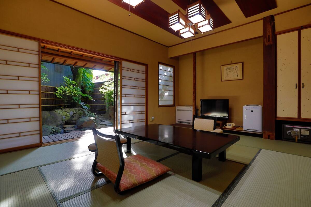 Ein traditioneller japanischer Raum mit Tatami-Boden, einem niedrigen Tisch, einem Stuhl, einem Fernseher und Blick auf einen Garten durch eine Schiebetür.
