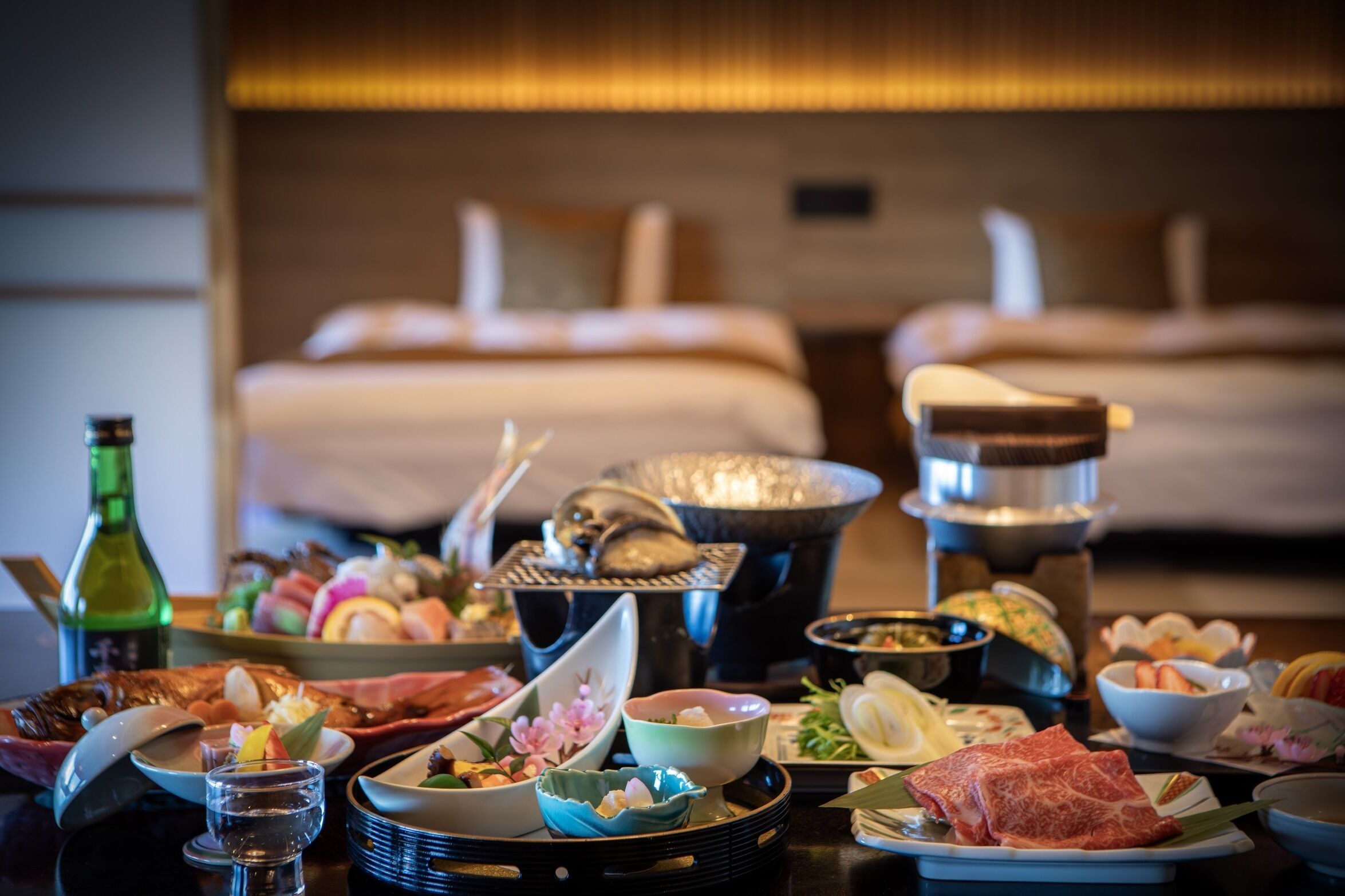 A dining table with various dishes and a hot pot, a bottle, and a bed in the background.